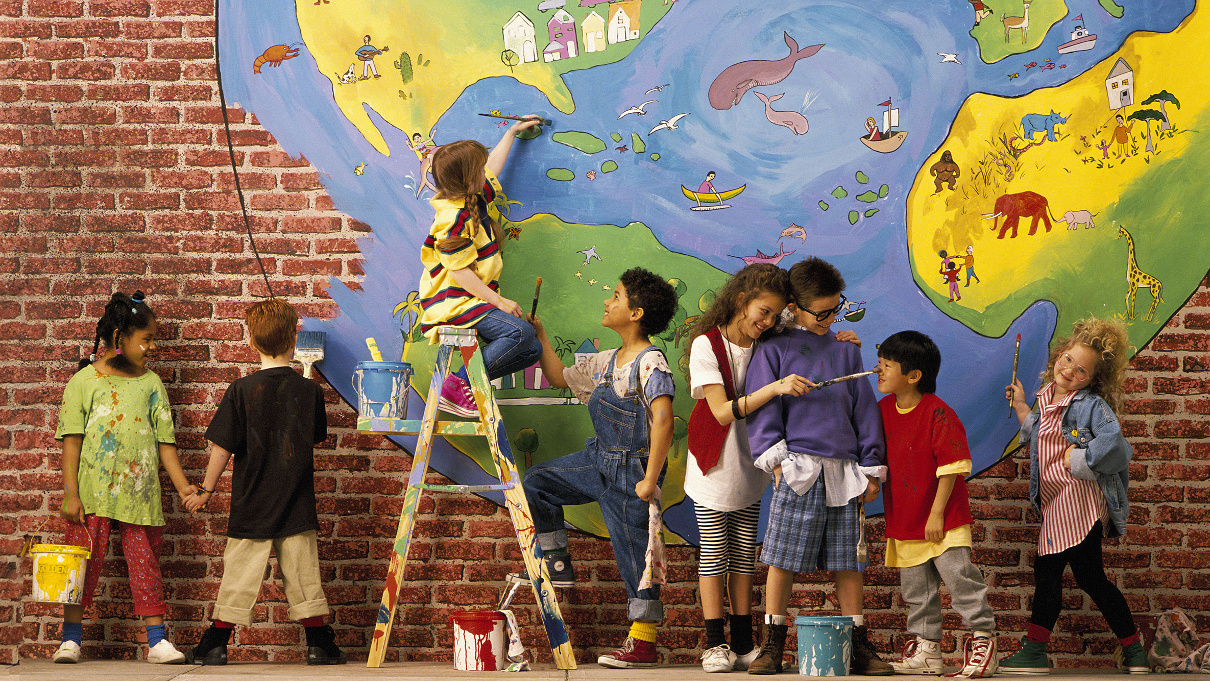 Children in colorful clothes paint a mural, and themselves a bit, on a brick wall.